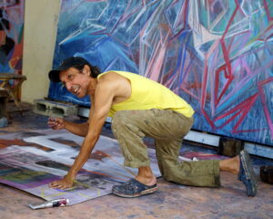 Artist Carlos Quintana in a yellow tanktop and tan pants, working on the floor of an art studio with a large blue and purple artwork behind